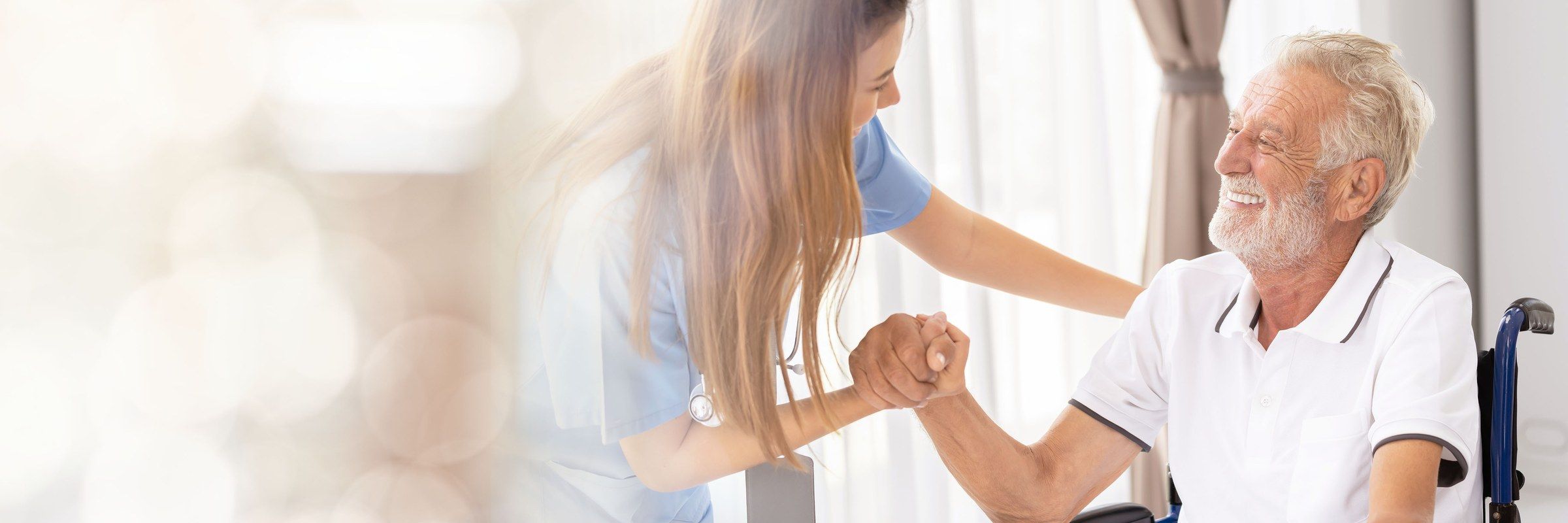 A nurse holds the hand of a smiling older man seated in a wheelchair by a sunlit window.