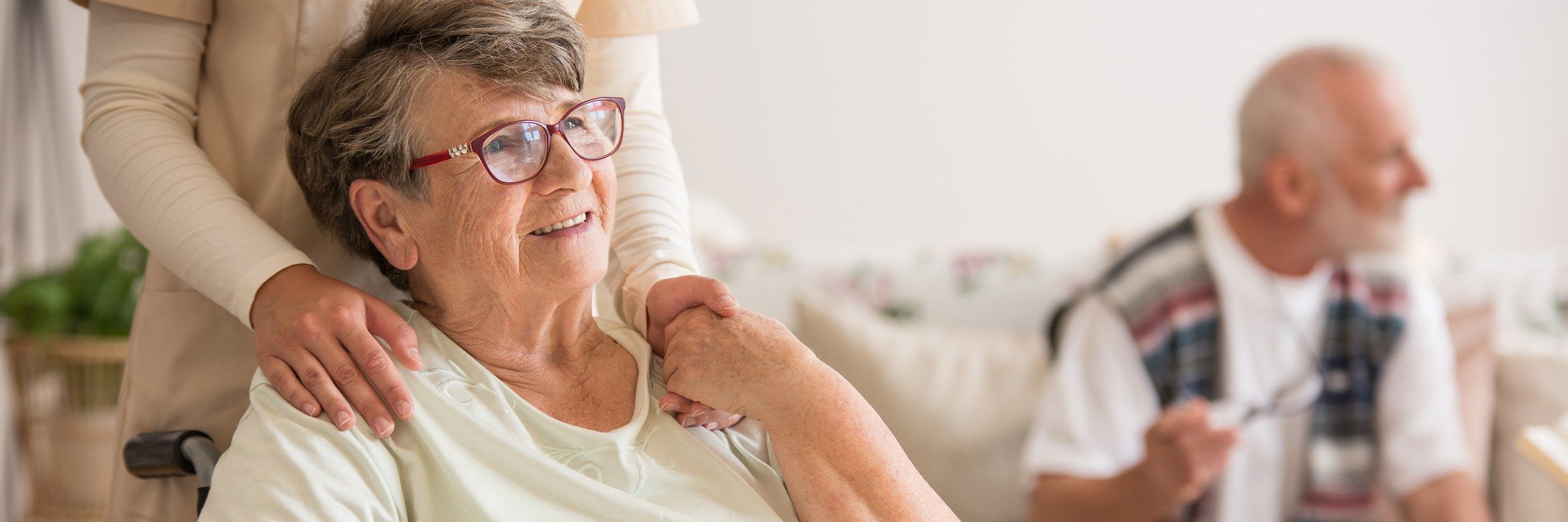 An older woman in glasses smiles while a caregiver rests both hands on her shoulders.