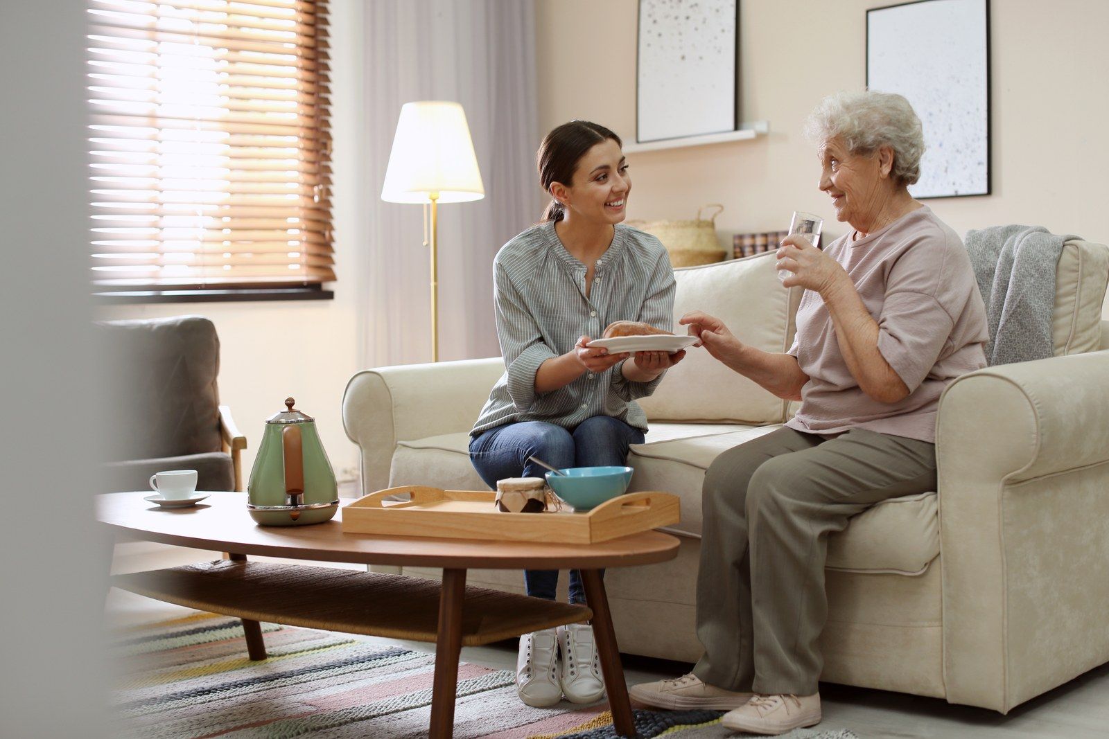 A visitor sits with an older woman in her living room, passing her a plate over a tea tray.