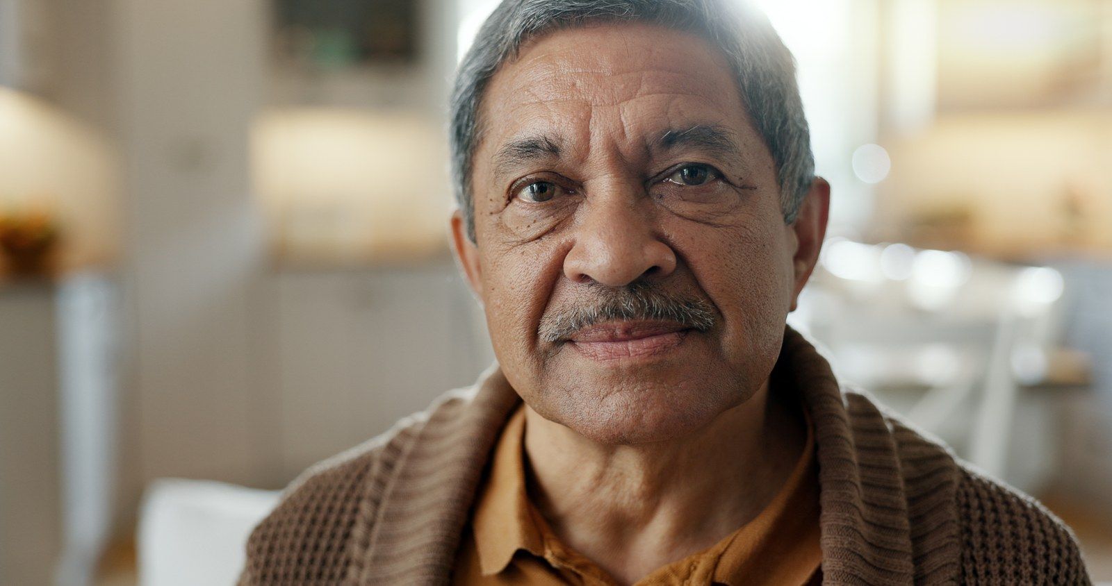 An older man in a brown cardigan at home, looking at the camera.